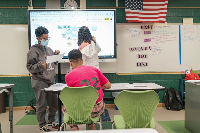 Three students work at a SMART Board in a classroom, with two standing and interacting with the display, while one sits at a desk in a bright pink shirt, focusing on their work.