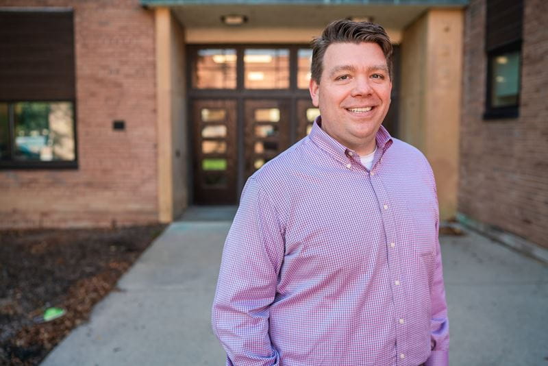 A man in a purple checkered shirt smiles while standing outside a brick school building, with double doors and windows behind him.