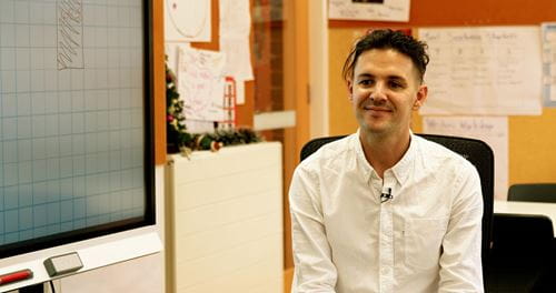 A male teacher in a white shirt sits near a SMART Board, sharing insights during a classroom discussion.