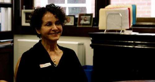 A school administrator smiles while seated in an office with framed photos and certificates in the background.