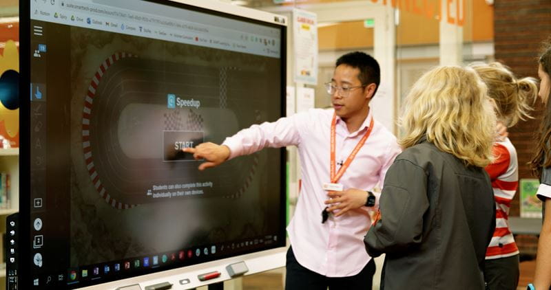 A teacher in a pink shirt points to an interactive activity on a SMART Board while students watch attentively.