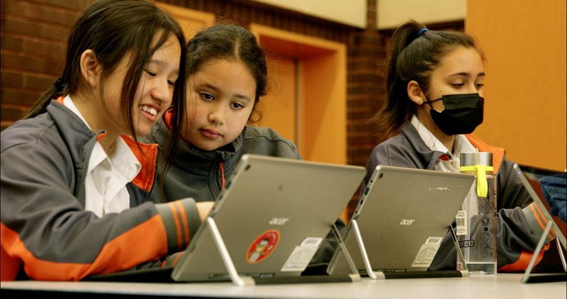 Three students collaborate on laptops during a classroom activity, with one wearing a mask.