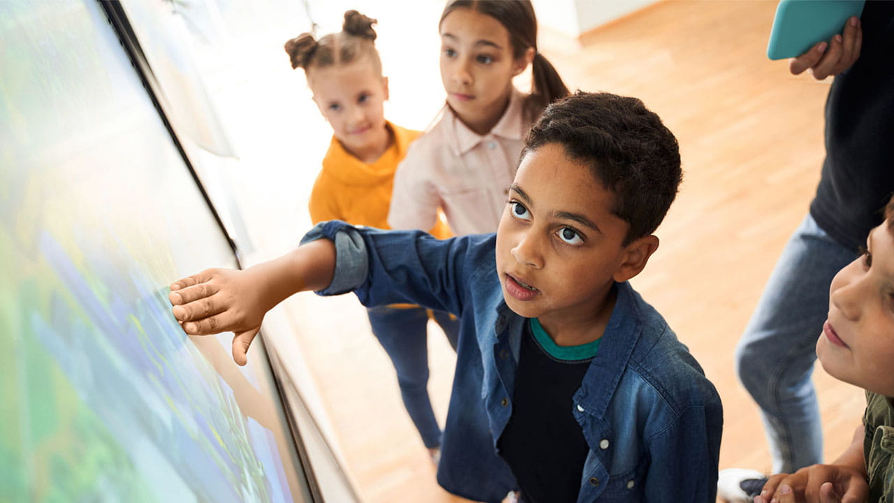 Primary aged students gathered around, and looking up at an interactive display. In the center of the image, a young boy looking amazed at the content on the screen.