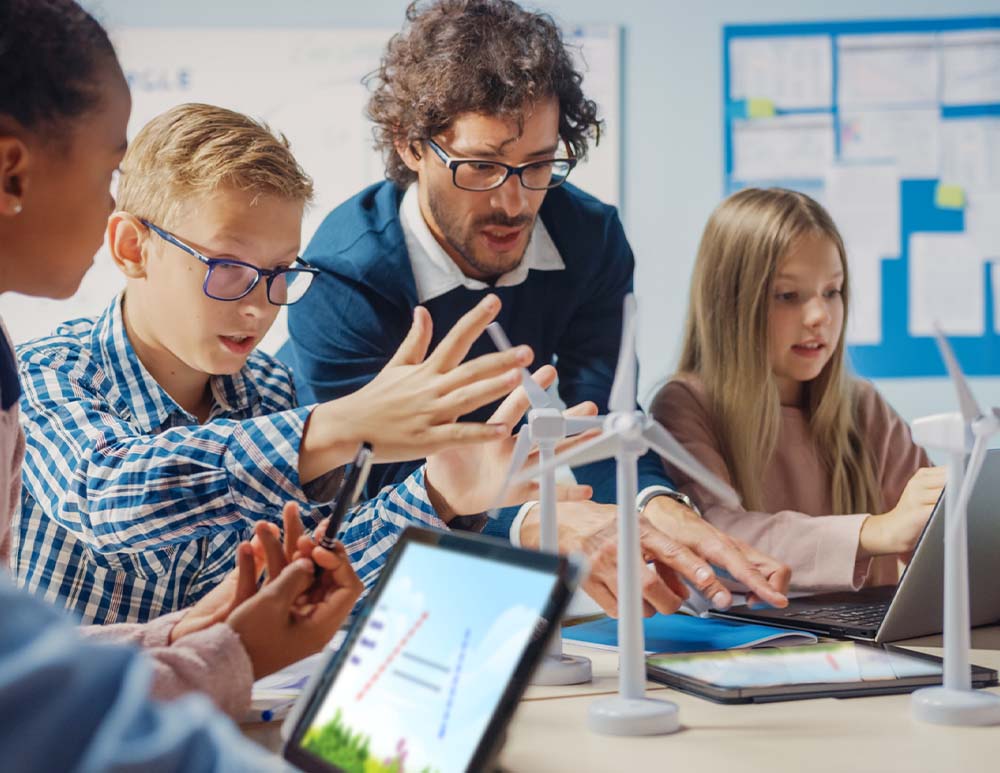 Teacher and students actively engaged with educational technology, including laptops and a wind turbine model, in a collaborative classroom setting.