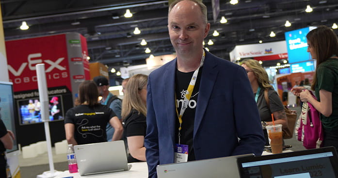 Lumio representative at a tech event, smiling while standing near laptops, with event attendees and booths in the background.