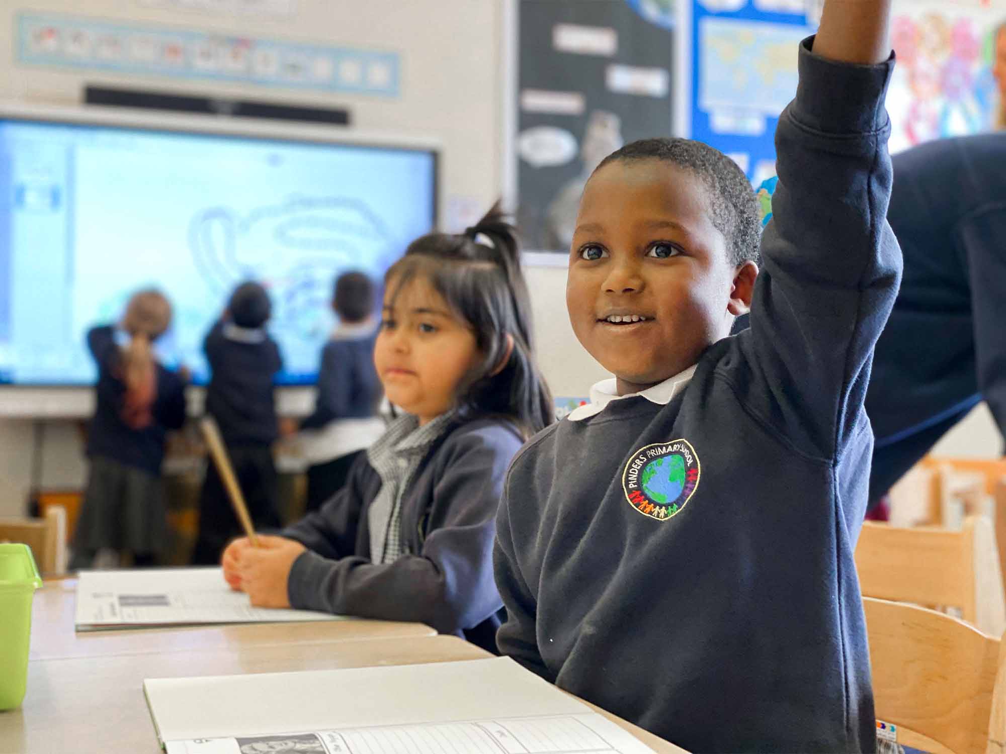 A young student sitting at a desk in a classroom, raising their hand to participate in class.