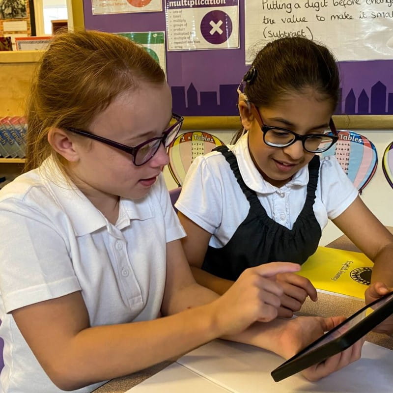 Two young students, focused on a tablet while seated in a classroom.
