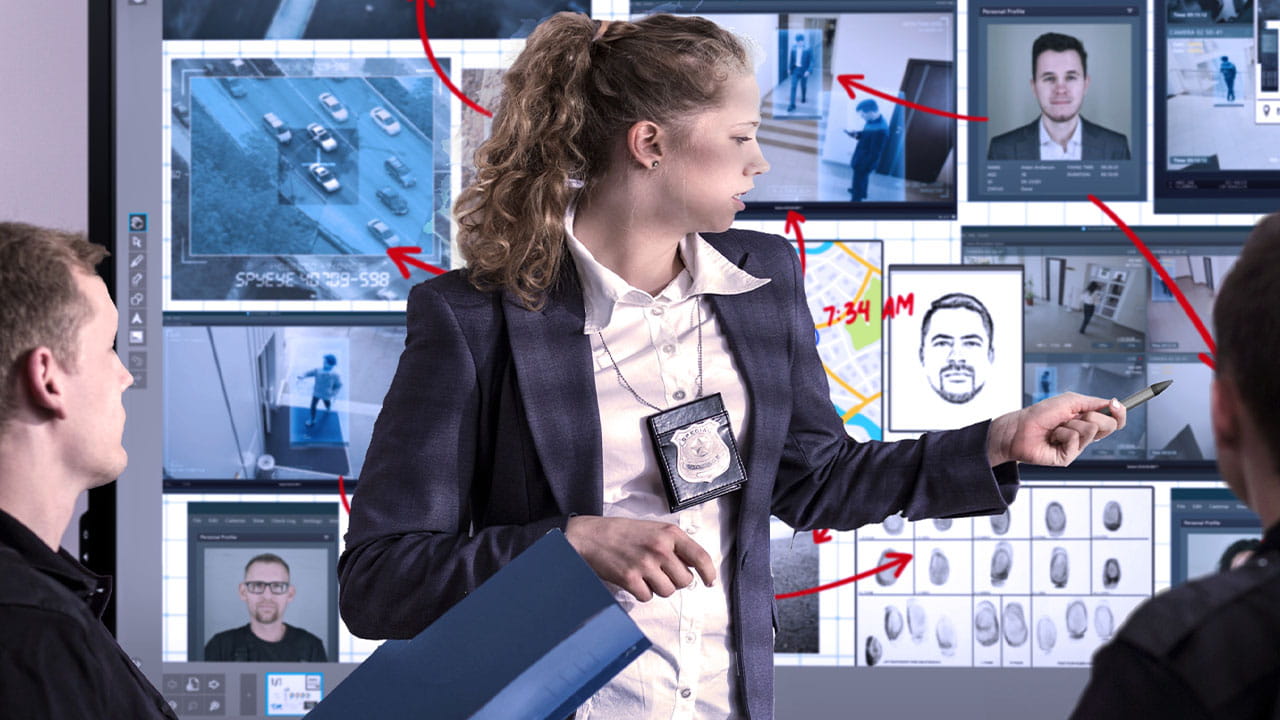 A female investigator points at a SMART interactive display showing multiple surveillance feeds, including car surveillance and suspect tracking, during a strategic discussion.