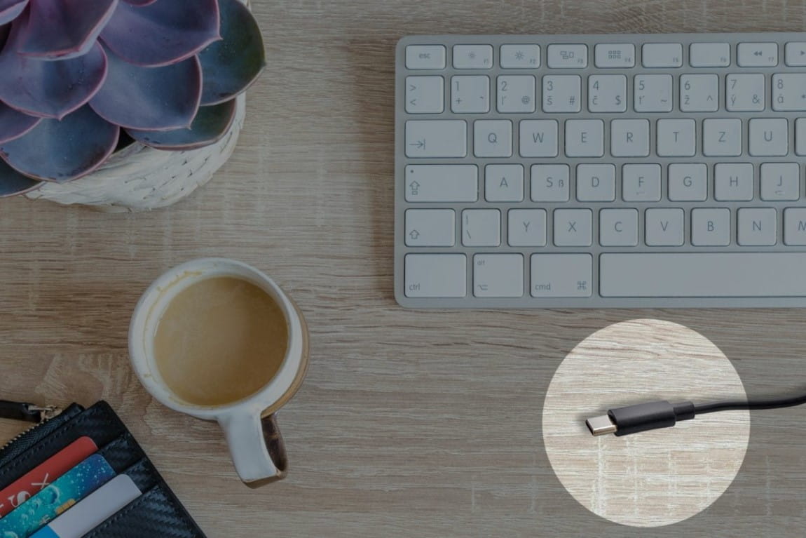 Close-up view of the USB-C cable on a wooden table, with a keyboard, coffee cup, and office supplies in a casual work environment.