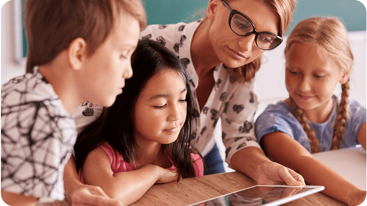 Elementary school teacher guiding students through a learning activity on a digital tablet in a classroom environment.